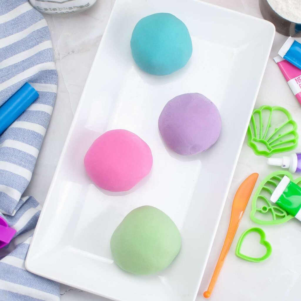 A white plate with a variety of colored homemade playdough balls and utensils.