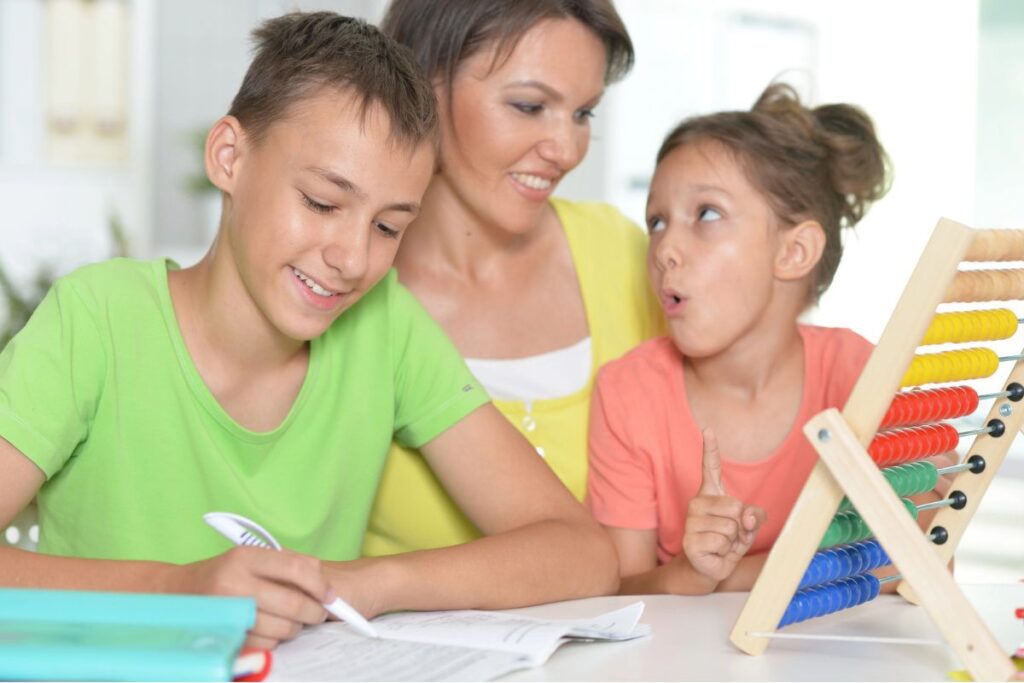 A woman sits between a boy writing in a notebook and a girl using an abacus. The boy is smiling, and the girl appears to be explaining something.