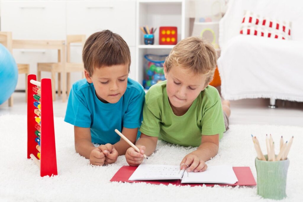 Two young boys lie on a white carpet, using colored pencils to write in a notebook. An abacus and a cup of pencils are nearby in a bright, organized room.