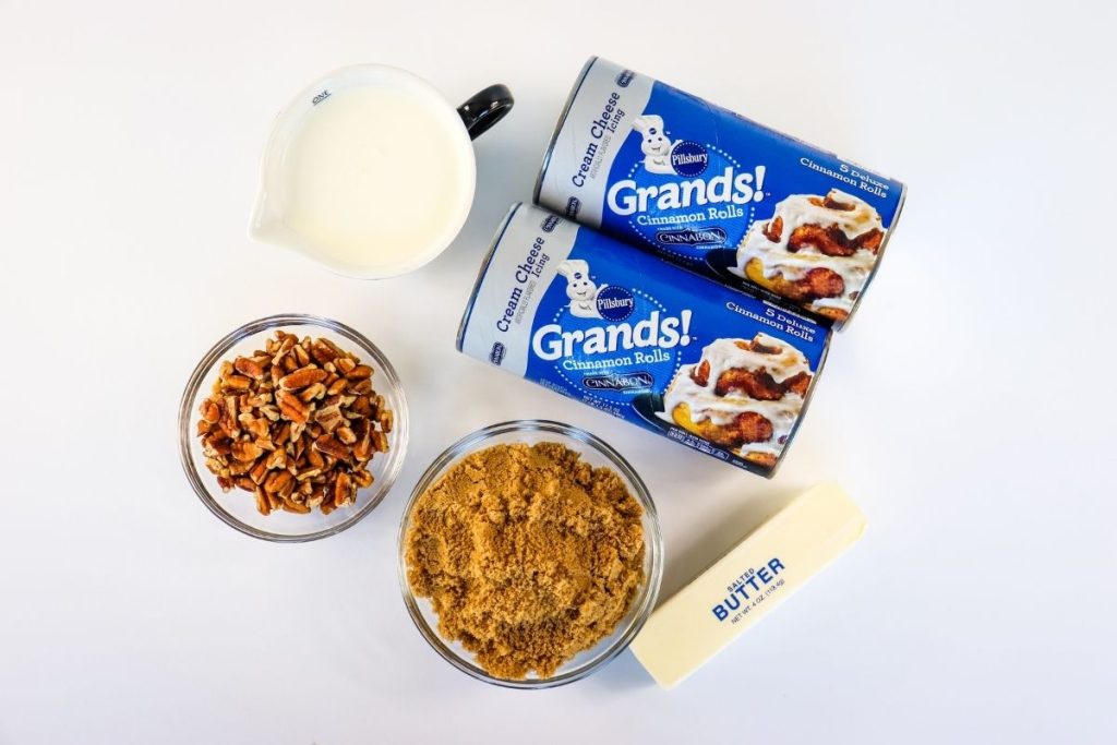 A pitcher of milk, two cans of Pillsbury Grands! cinnamon rolls, a bowl of chopped pecans, a bowl of brown sugar, and a stick of butter on a white surface.