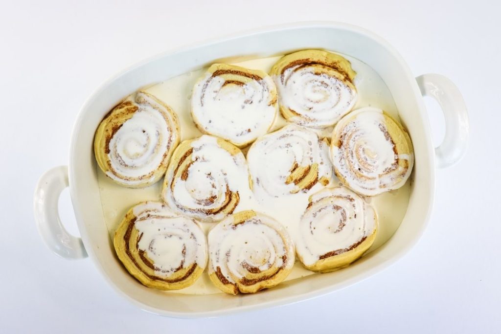 A white baking dish containing nine cinnamon rolls covered with a layer of white icing, arranged on a white surface.
