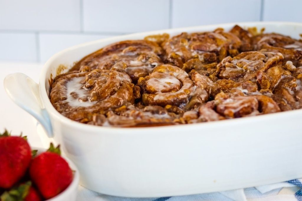 A white baking dish filled with glazed cinnamon rolls sits on a counter, with a bowl of fresh strawberries nearby.
