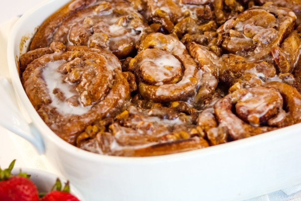 A white baking dish filled with butter pecan cinnamon rolls, partly covered in icing; fresh strawberries are visible in the foreground.