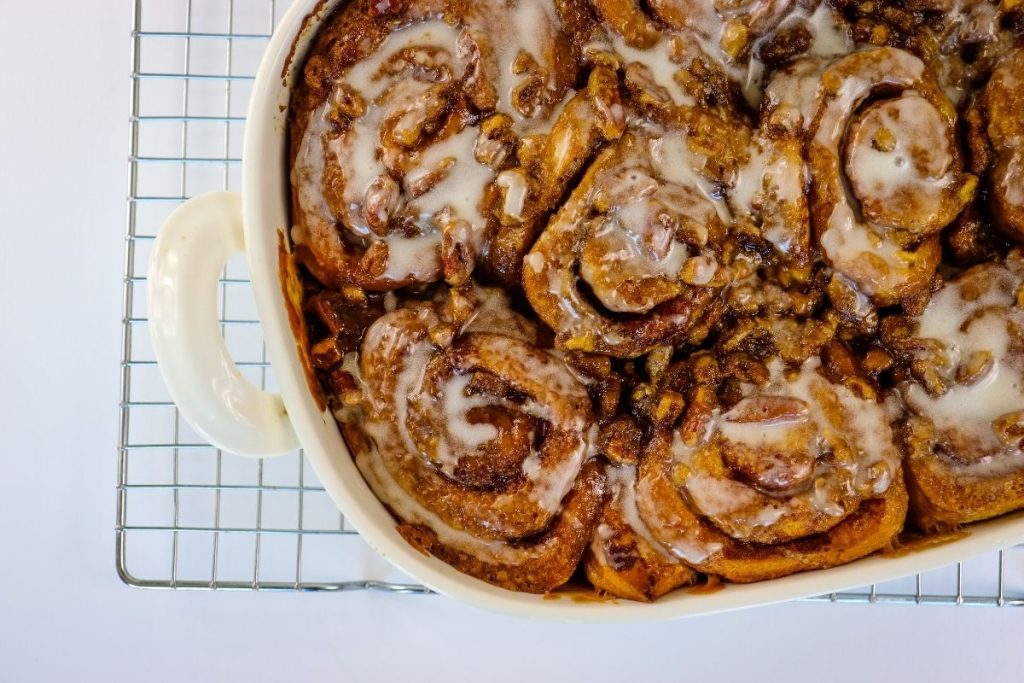 A baking dish filled with iced cinnamon rolls topped with chopped nuts sits on a cooling rack against a white background.