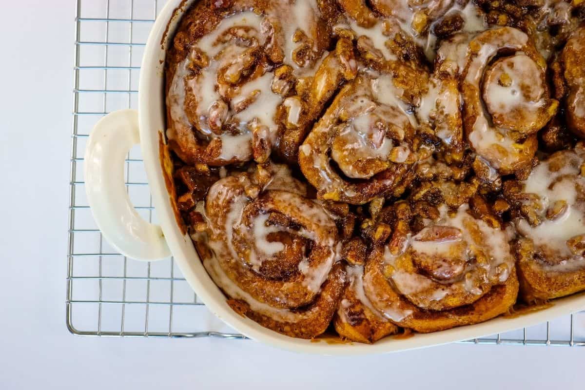 A baking dish filled with iced cinnamon rolls topped with chopped nuts sits on a cooling rack against a white background.