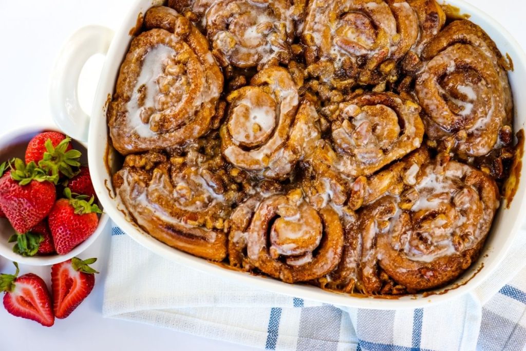 A baking dish filled with butter pecan cinnamon rolls sits next to a bowl of fresh strawberries on a white and blue towel.