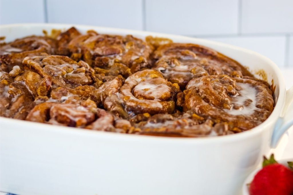 A white baking dish filled with glazed cinnamon rolls sits on a countertop with a strawberry in the corner of the frame.