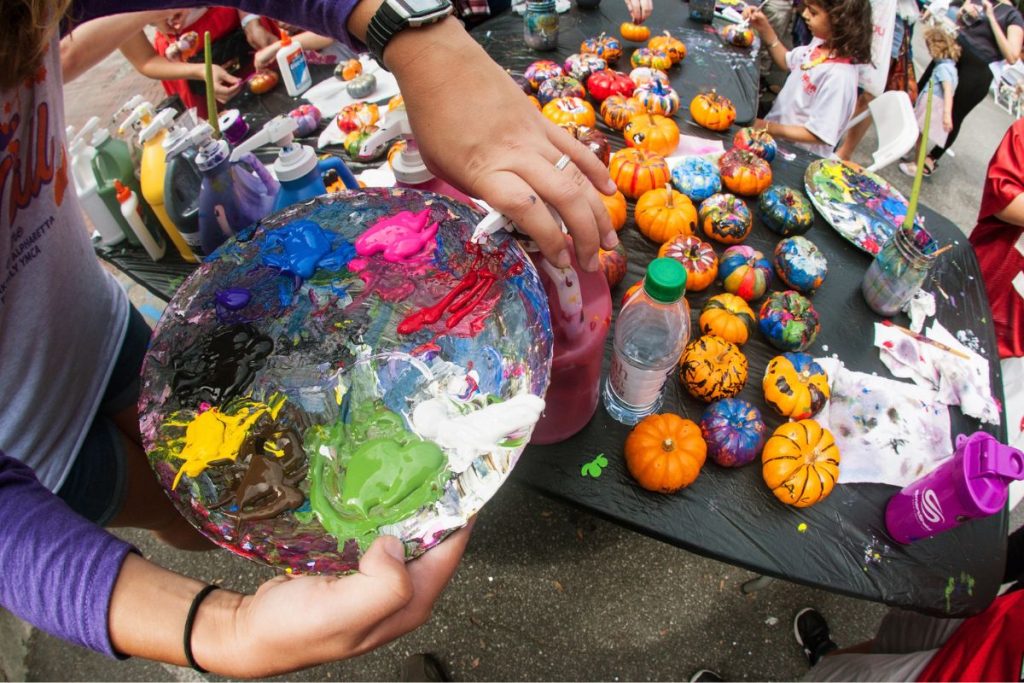 A person holds a paint palette near a table covered with small painted pumpkins, bottles of paint, and water, with people participating in a pumpkin decorating activity.