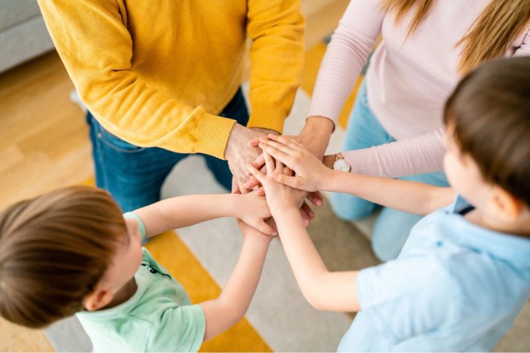 Four people, two adults and two children, stand in a circle and stack their hands together in the center of the group.