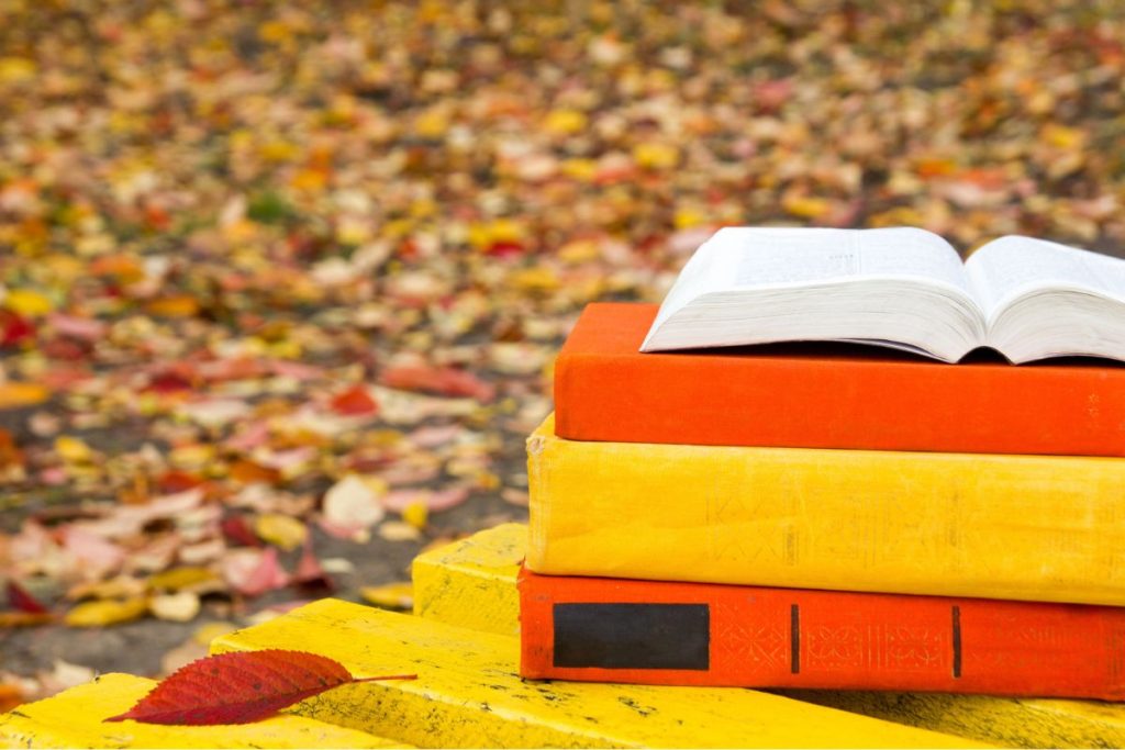 Three books, one open, are stacked on a yellow bench outdoors with autumn leaves scattered on the ground in the background.