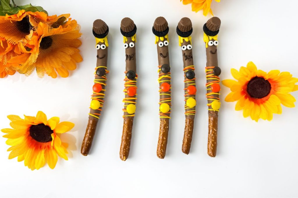 Five decorated pretzel sticks resembling whimsical figures are surrounded by artificial yellow sunflowers on a white surface.