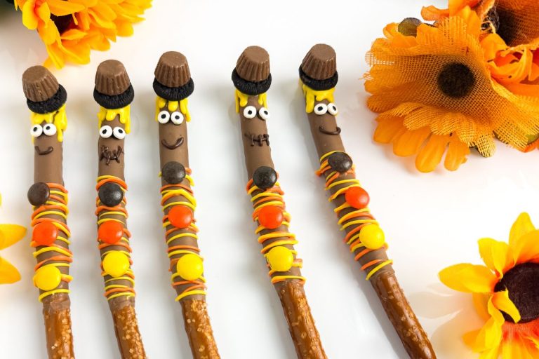 Five pretzel sticks decorated as turkeys with candy eyes, peanut butter cups, chocolate hats, and colorful candy, arranged among artificial yellow-orange sunflowers.