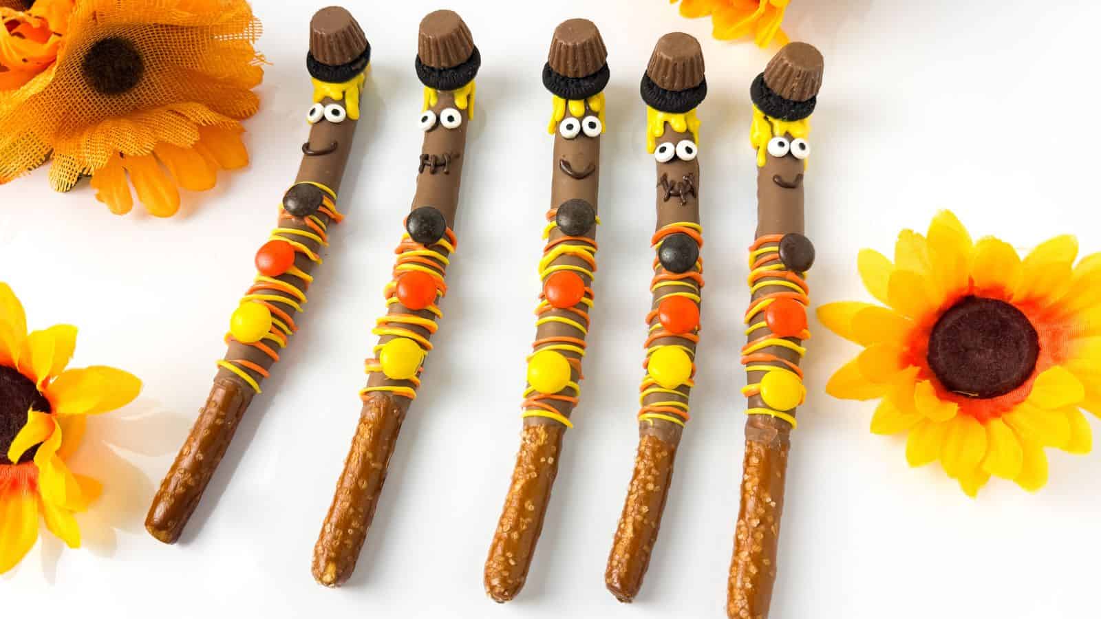 Five pretzel sticks are decorated with yellow, orange, and brown candy to resemble scarecrows, with candy hats and faces, surrounded by artificial sunflowers on a white surface.