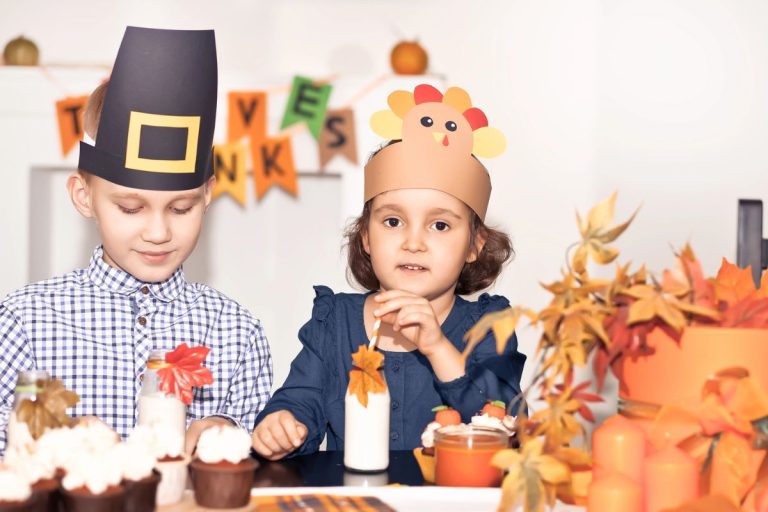 Two children wearing Thanksgiving-themed hats sit at a decorated table with cupcakes, milk bottles, and autumn leaves. A "Thanks" banner is visible in the background.