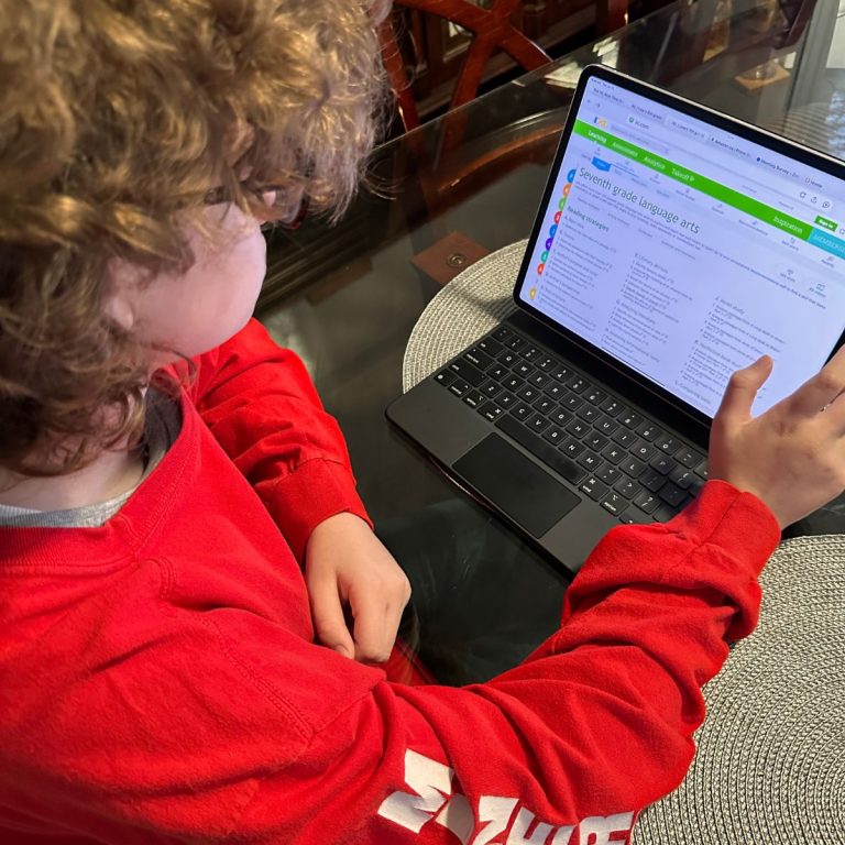 A child in a red shirt uses a laptop at a table, viewing IXL as part of her summer learning