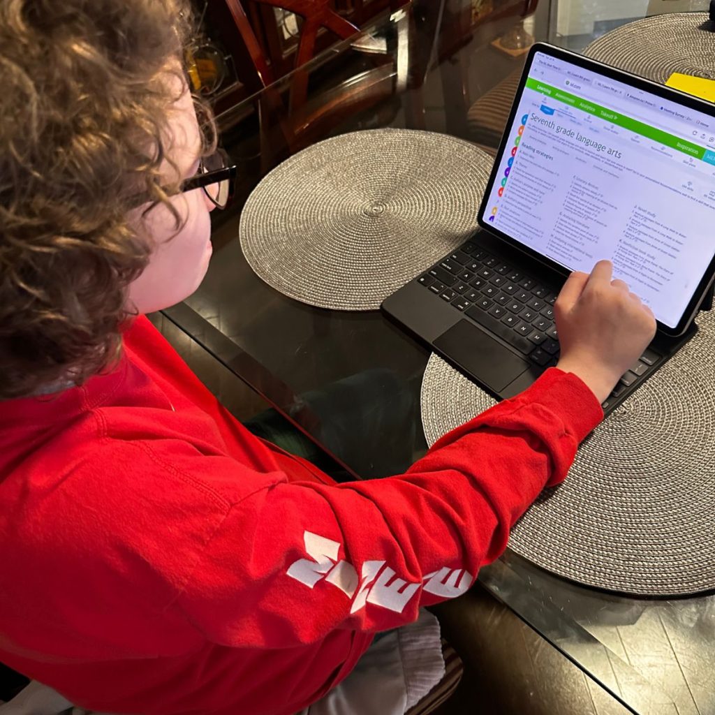 A person in a red long-sleeve shirt uses a touchscreen laptop at a table, viewing an IXL homeschool webpage with a list of English language arts resources.