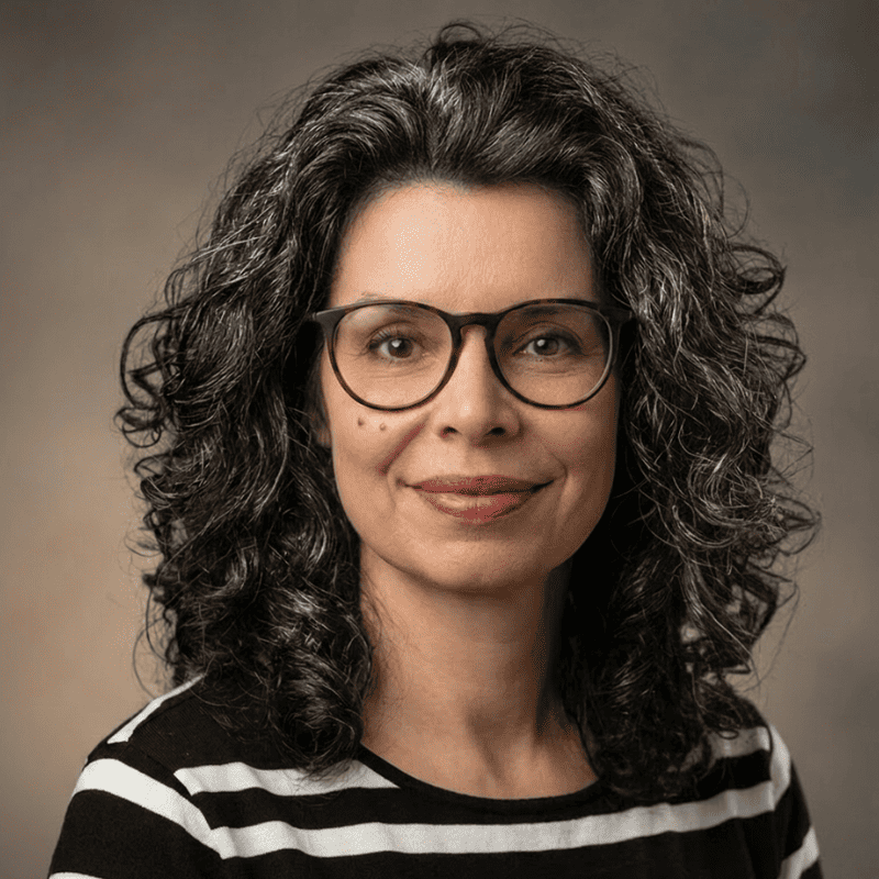 A woman with curly dark hair and glasses, wearing a black and white striped top, smiles slightly in a studio portrait against a plain background.